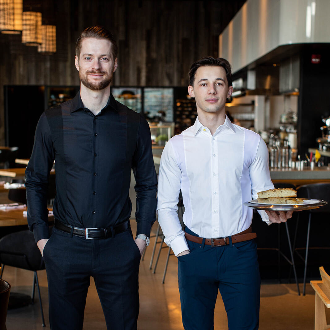 Two men wearing slim fit Men's Work Shirt in black and white, standing in a modern restaurant setting.