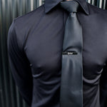 Close-up of a man wearing a black Never-Iron Work Shirt with a black tie and tie clip against a metal background