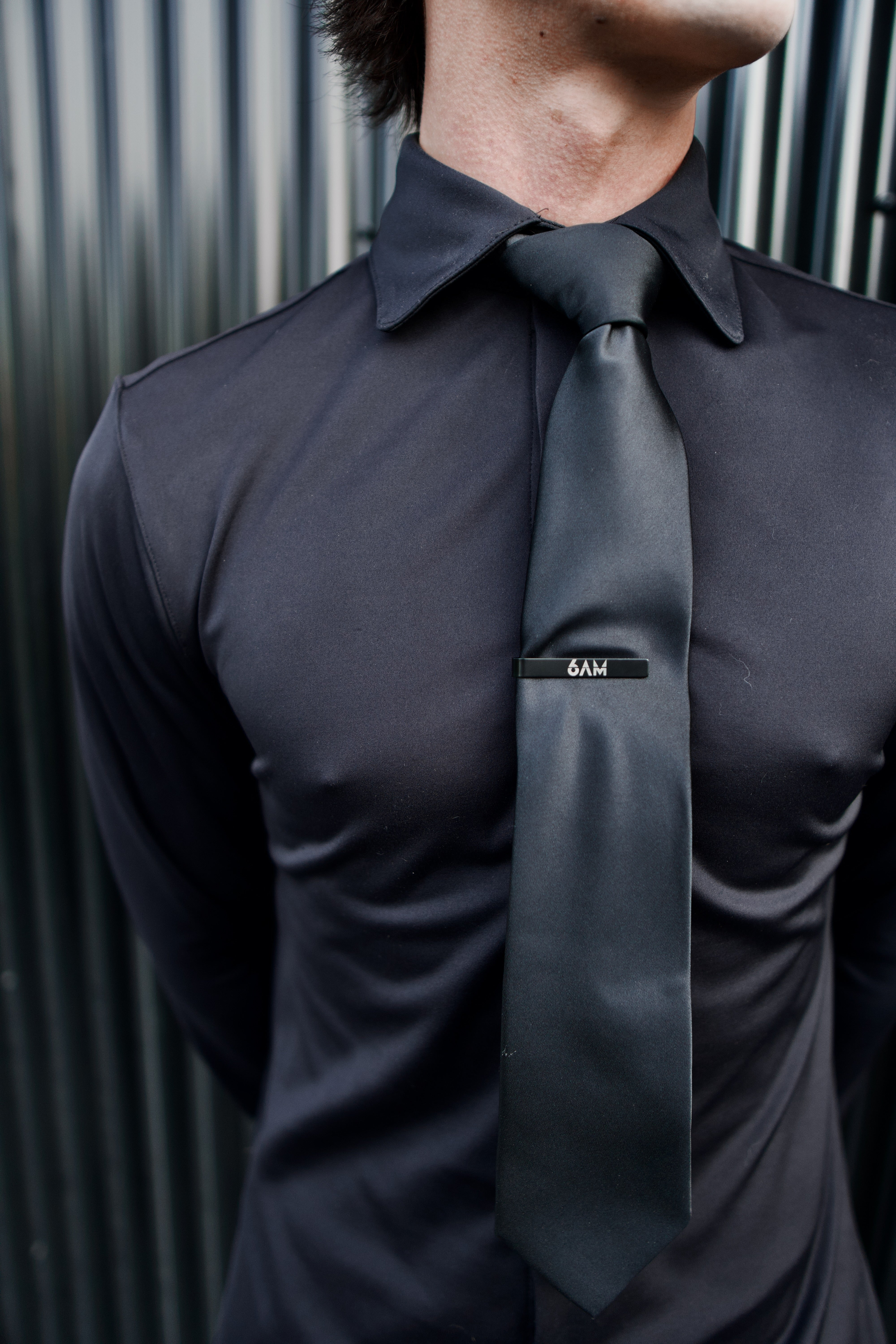 Close-up of a man wearing a black Never-Iron Work Shirt with a black tie and tie clip against a metal background