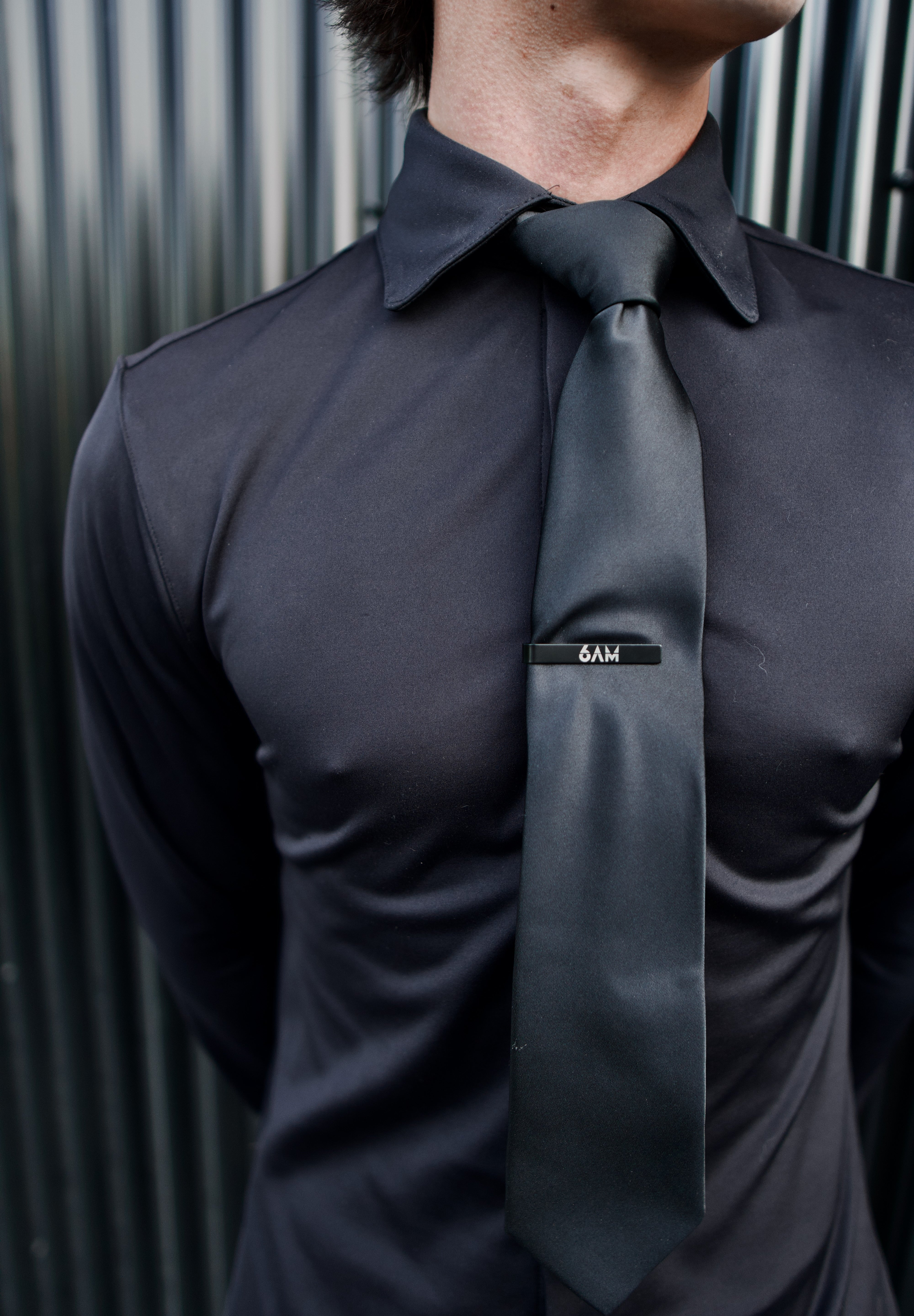 Close-up of a man wearing a black Never-Iron Work Shirt with a black tie and tie clip against a metal background