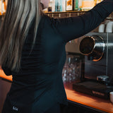 Woman wearing black Women's Work Shirt reaching for a bottle behind a bar counter with coffee machine nearby