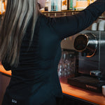 Woman wearing black Women's Work Shirt reaching for a bottle behind a bar counter with coffee machine nearby