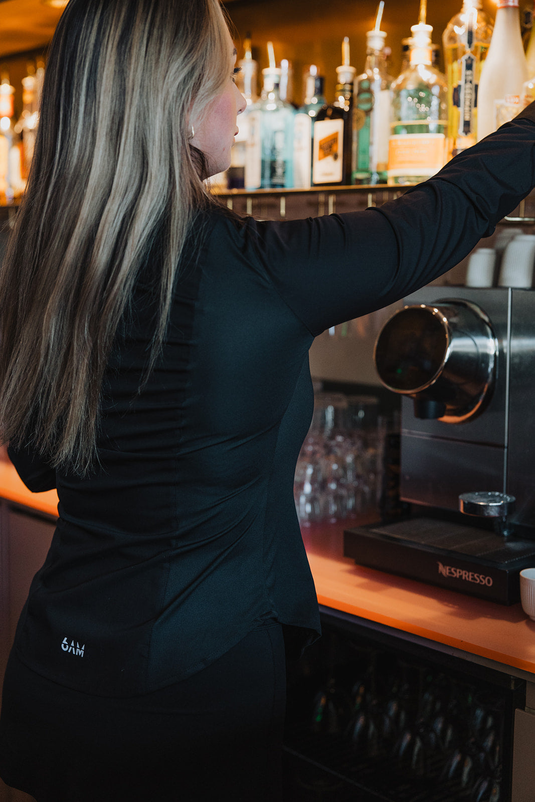 Woman wearing black Women's Work Shirt reaching for a bottle behind a bar counter with coffee machine nearby