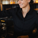 Woman wearing black Women's Work Shirt smiling while working behind a counter in a cozy setting