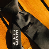 6AM Formal Black Tie close-up on wooden surface featuring black fabric and 6AM logo on the tail.