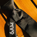 6AM Formal Black Tie close-up on wooden surface featuring black fabric and 6AM logo on the tail.