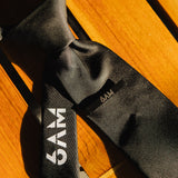 6AM Formal Black Tie close-up on wooden surface featuring black fabric and 6AM logo on the tail.